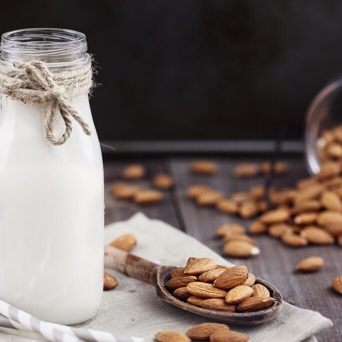 Organic white almond milk in a glass bottle with whole almonds spilled over a rustic wooden table.