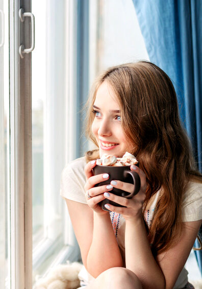 Beautiful young woman drinking coffee and looking through window while sitting at windowsill at home